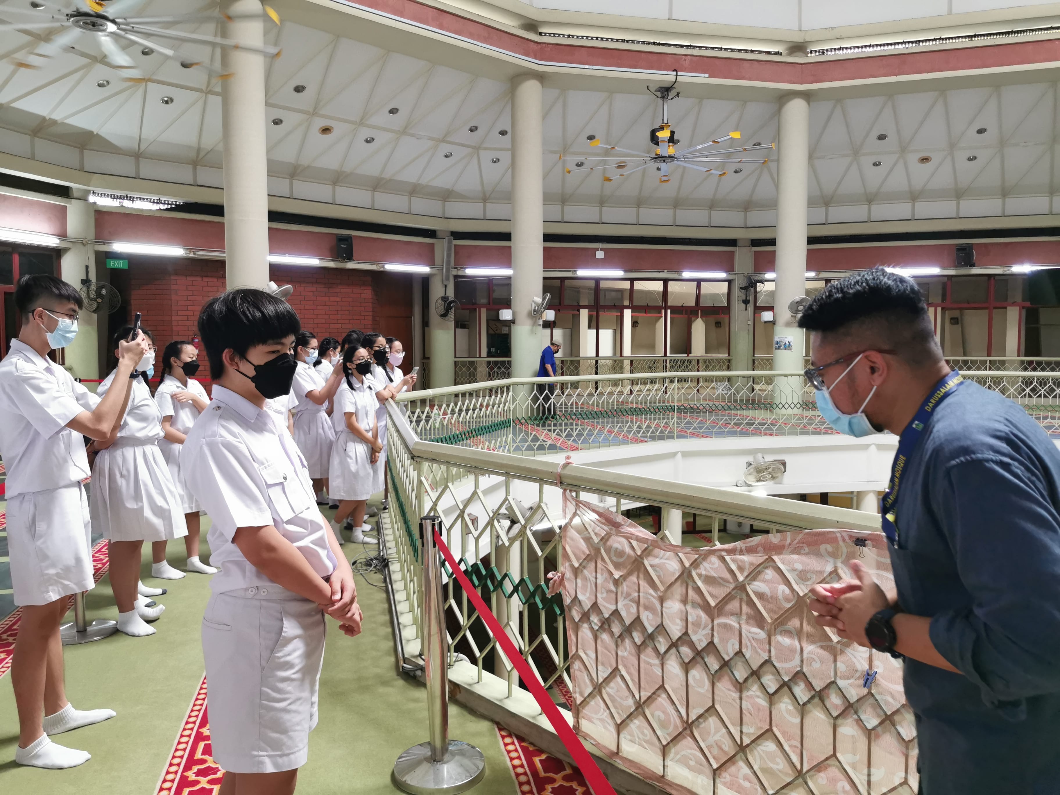 Students in white uniforms wearing masks stand on a balcony inside a mosque, listening to a guide.